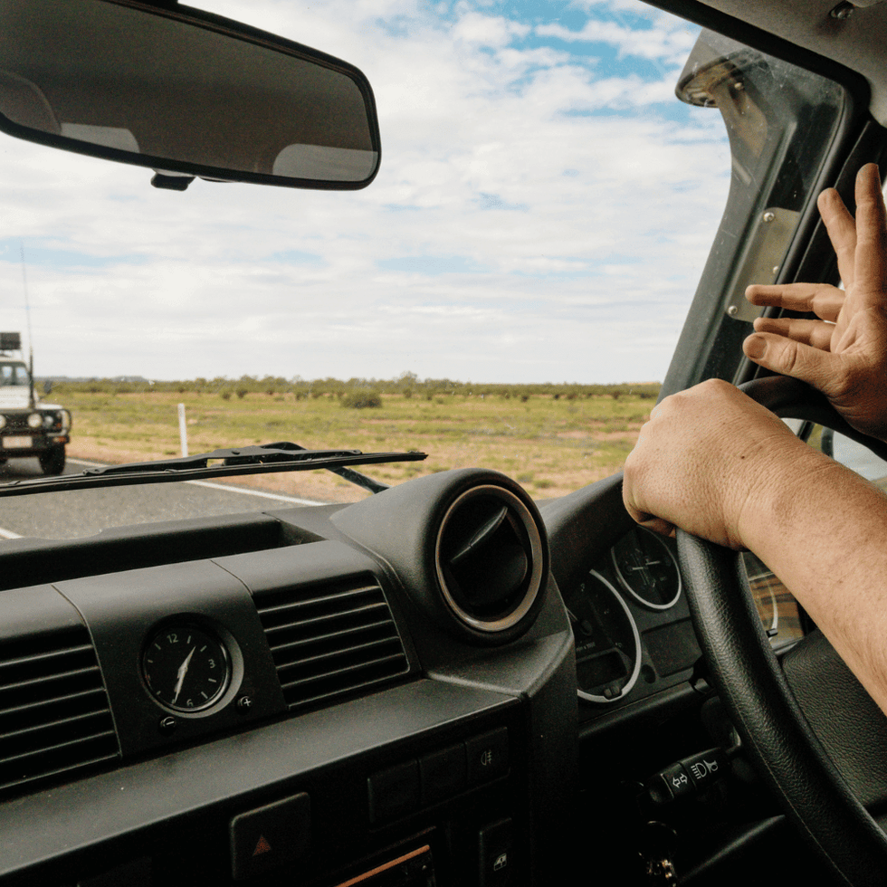 Driver inside a 4WD vehicle on an open road, signaling with two fingers while passing another off-road vehicle in the Australian outback.