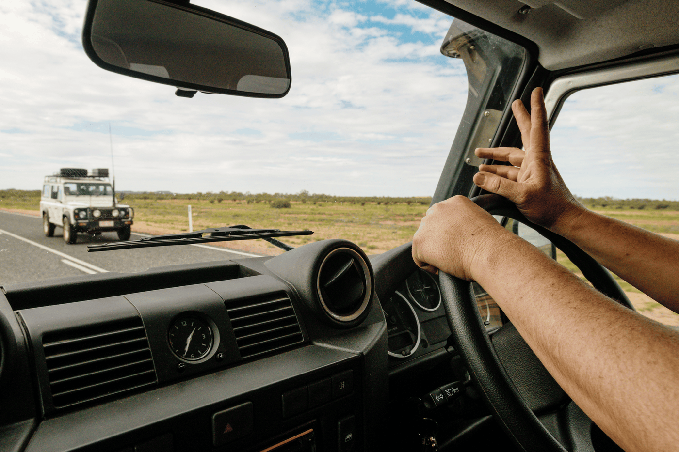 Driver inside a 4WD vehicle on an open road, signaling with two fingers while passing another off-road vehicle in the Australian outback.