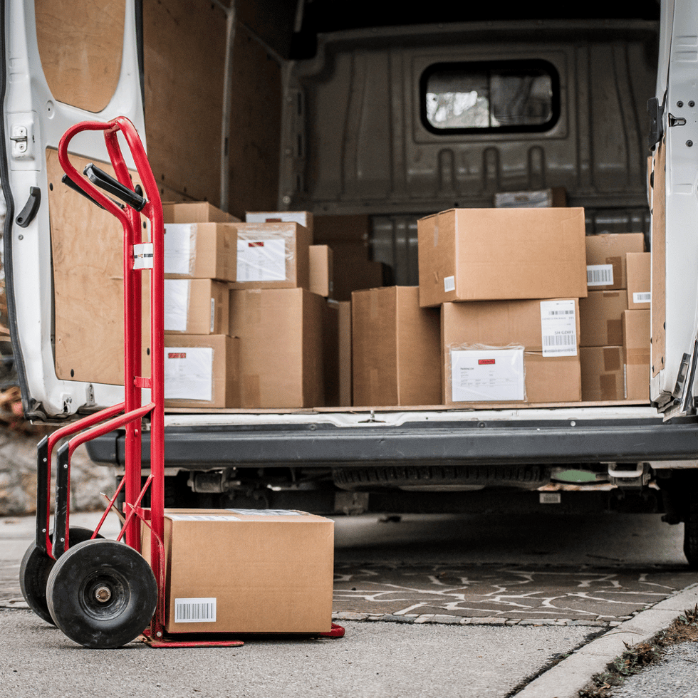 Open delivery van loaded with cardboard boxes, featuring a red dolly cart in the foreground ready for unloading