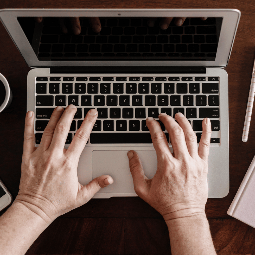 Person typing on a laptop with a cup of coffee, smartphone displaying an image, and a pen and notebook on a wooden desk.