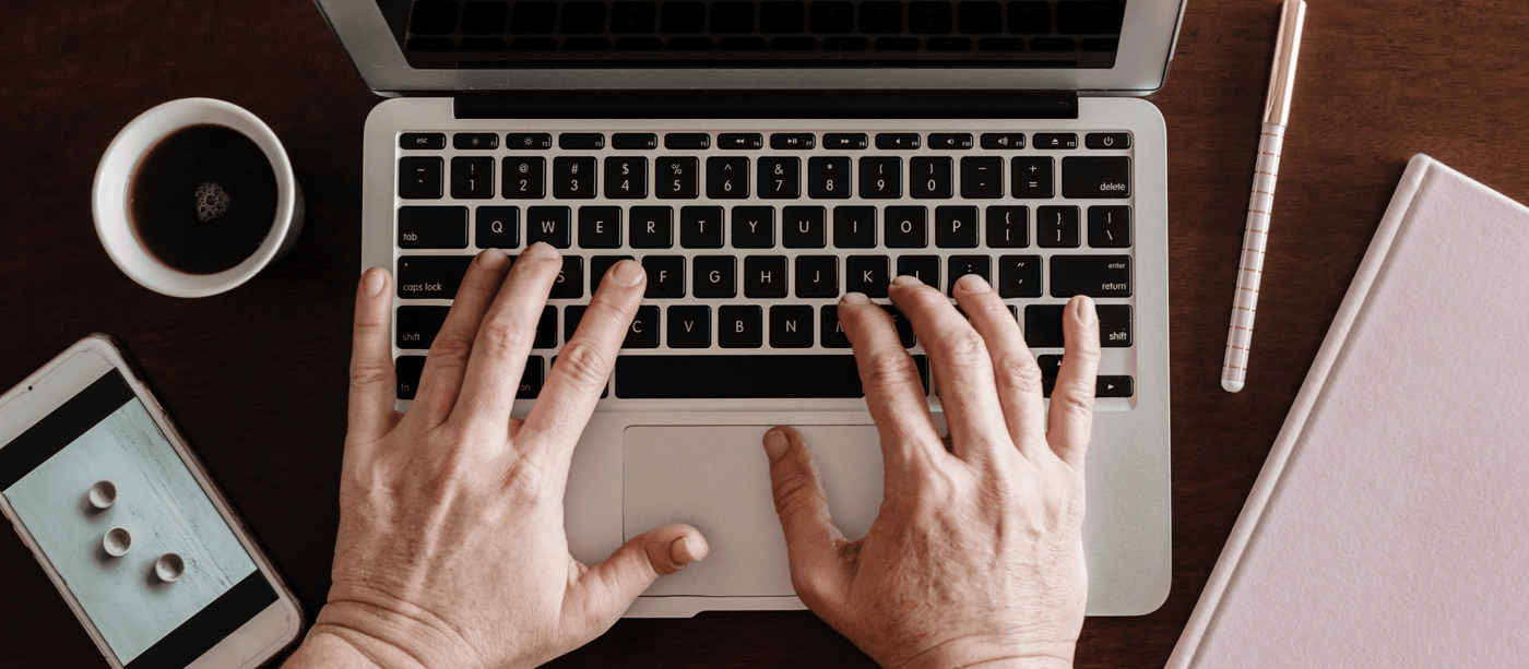 Person typing on a laptop with a cup of coffee, smartphone displaying an image, and a pen and notebook on a wooden desk.