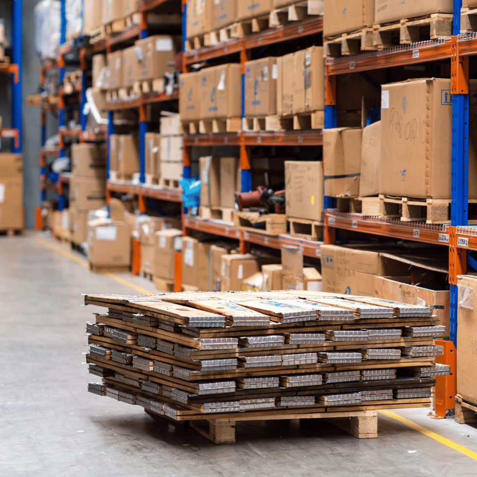 Interior of a large warehouse with stacked cardboard boxes on pallets, organized on orange and blue shelving racks, and a stack of pallets in the foreground.