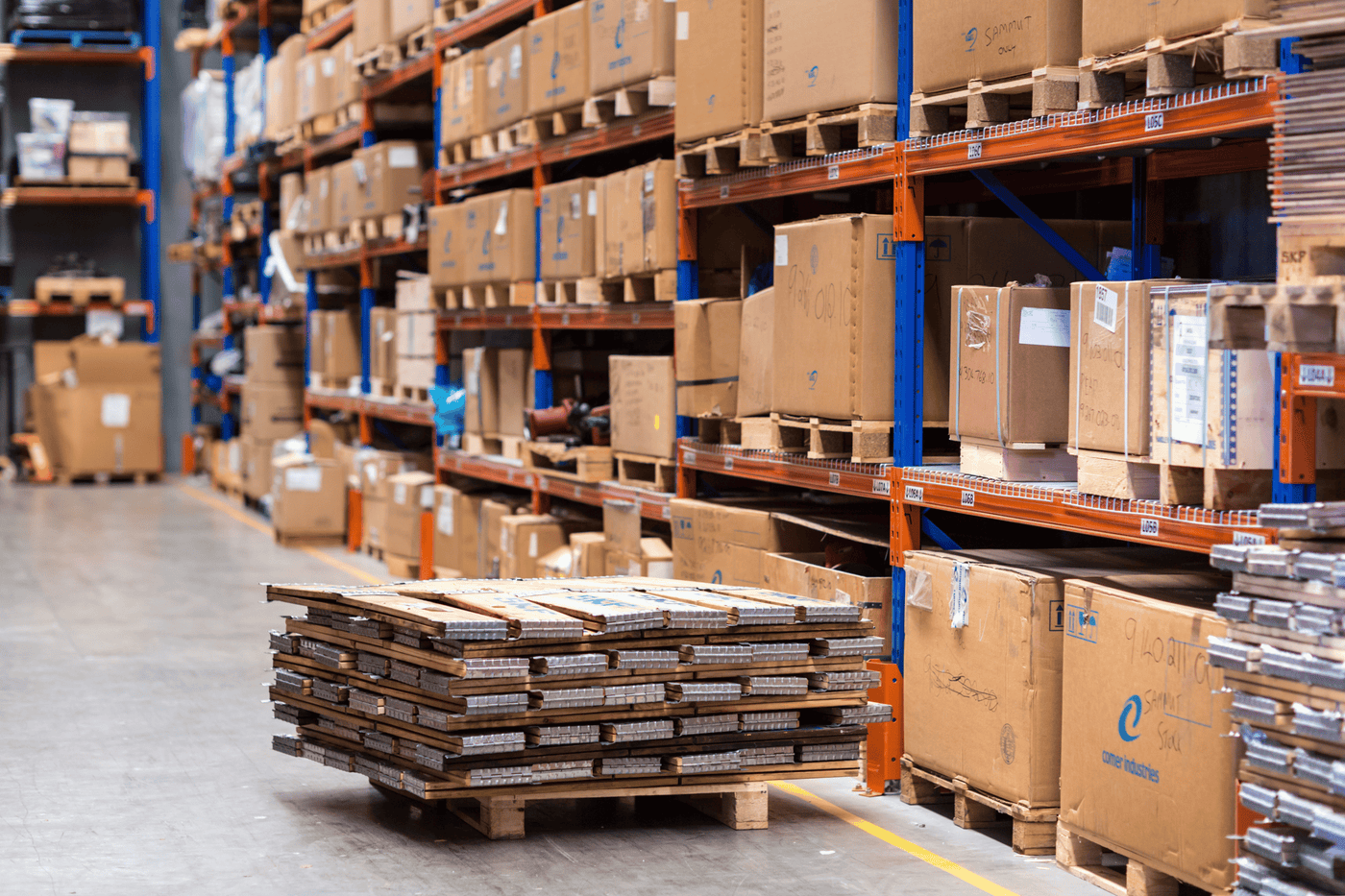 Interior of a large warehouse with stacked cardboard boxes on pallets, organized on orange and blue shelving racks, and a stack of pallets in the foreground.